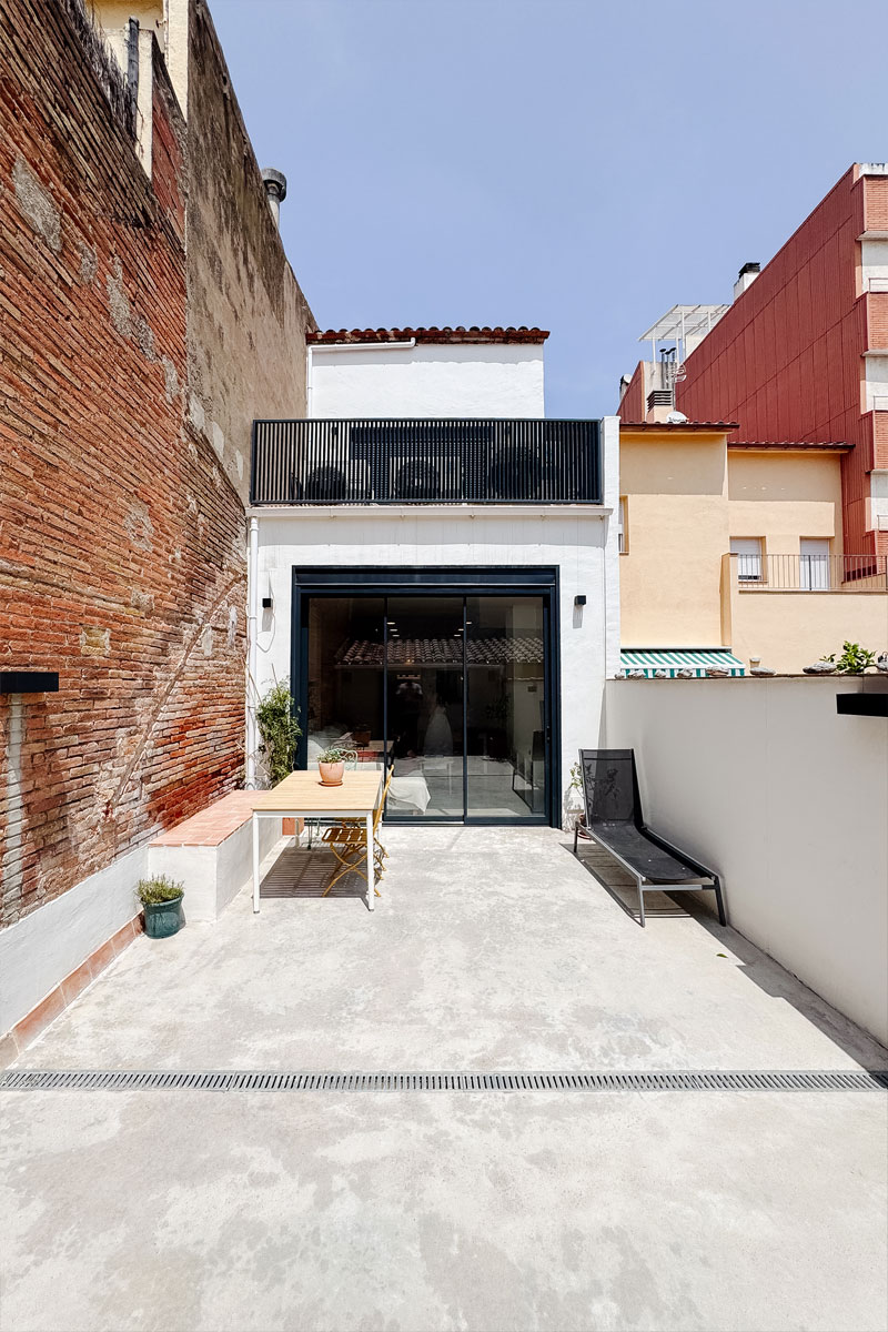 Patio reformado en Sabadell tras el cambio de uso, con pavimento de hormigón, mobiliario exterior y nueva fachada blanca con carpinterías negras y terraza