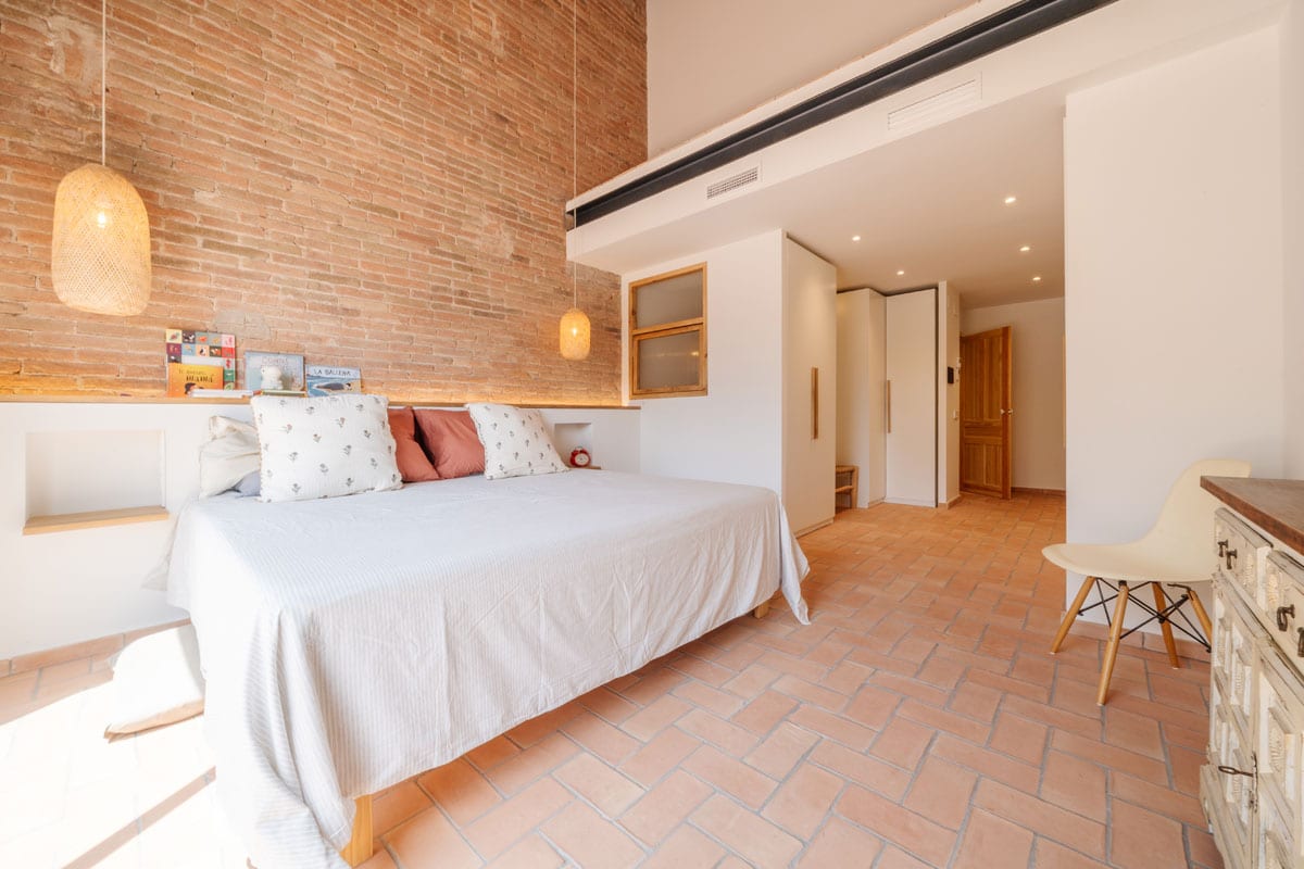 Dormitorio reformado en Sabadell tras el cambio de uso, con pared de ladrillo vista, pavimento de barro cocido y diseño interior cálido y acogedor