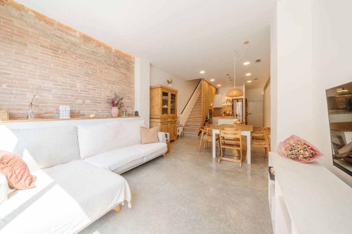 Salón comedor reformado en vivienda saludable de Sabadell, con pared de ladrillo visto, sofá blanco y cocina abierta al fondo