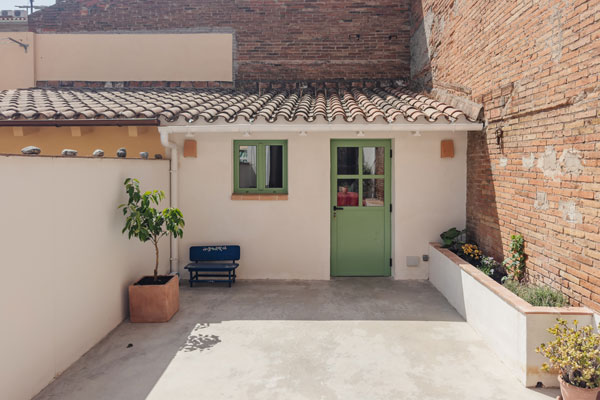 Patio en vivienda saludable de Sabadell, con fachada reformada en blanco roto, puerta y ventanas verdes y jardineras laterales con vegetación.