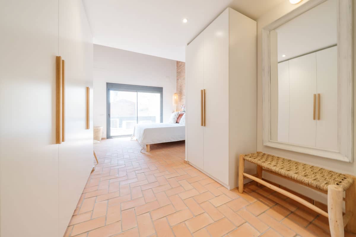 Vestidor conectado al dormitorio principal en vivienda saludable de Sabadell, con armarios empotrados blancos, banco de fibras naturales y espejo de cuerpo entero