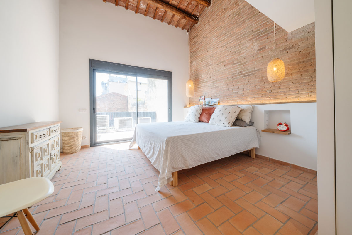 Habitación luminosa en vivienda saludable tras el cambio de uso en Sabadell, con pavimento de barro cocido, pared de ladrillo visto y mobiliario natural