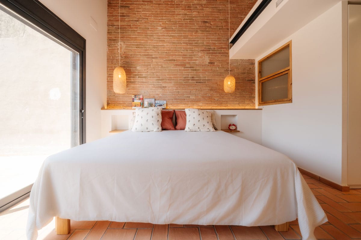 Dormitorio principal reformado en Sabadell, con pared de ladrillo visto, iluminación cálida y gran ventanal hacia el exterior