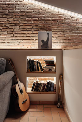 Rincón de lectura en vivienda reformada en Sabadell, con estanterías empotradas iluminadas, pared de ladrillo visto y guitarra apoyada en el sofá