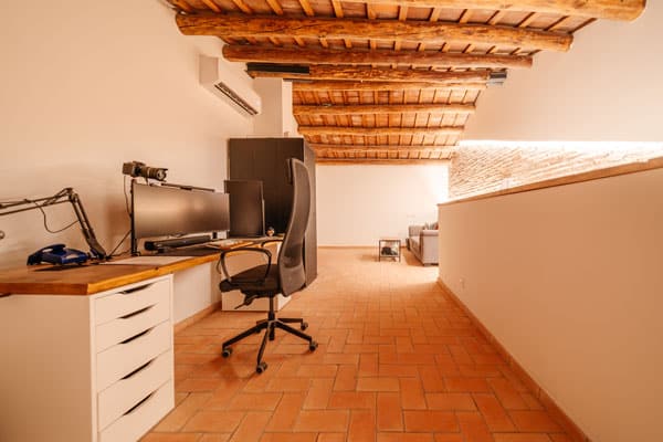 Despacho en altillo de vivienda saludable en Sabadell, con techo de vigas de madera, suelo de barro cocido y escritorio de trabajo con ordenador